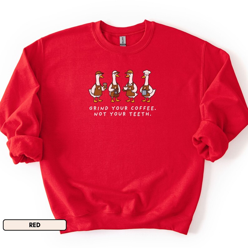 May include: A red crew neck sweatshirt. The front displays a graphic of four geese dressed as baristas, holding coffee cups and wearing aprons and hats. The text "GRIND YOUR COFFEE. NOT YOUR TEETH." is below the geese.