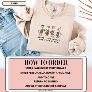 May include: A sand-coloured sweatshirt with a crew neck. The front displays a graphic of four cartoon geese holding coffee cups, with the text "GRIND YOUR COFFEE. NOT YOUR TEETH." The sweatshirt is shown with blue jeans.
