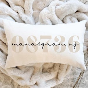 May include: Cream-colored rectangular pillow with the text "manasquan, nj" and the numbers "08736" printed on it in a neutral color. The pillow is resting on a soft, cream-colored blanket.