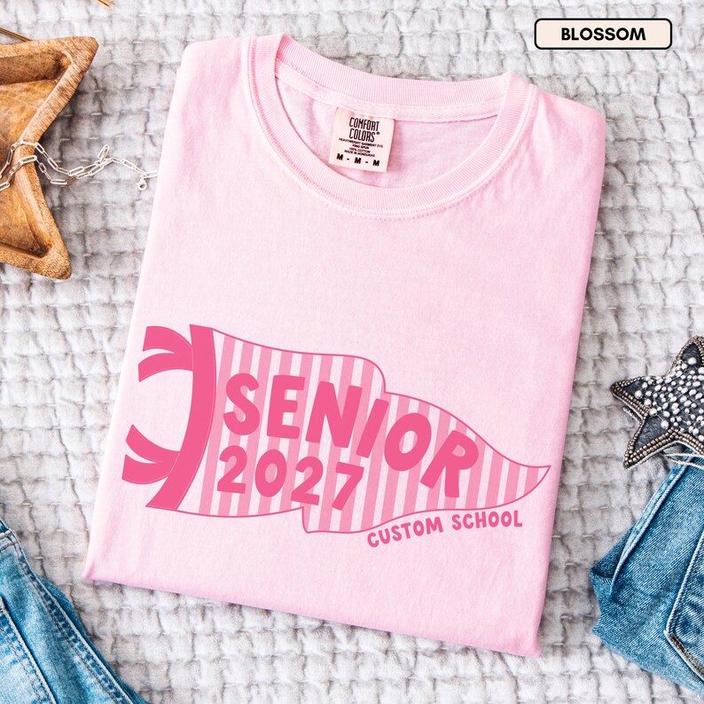 May include: A light pink t-shirt featuring a dark pink pennant graphic with vertical stripes. The text inside the pennant reads SENIOR 2027, and the text below reads CUSTOM SCHOOL. The shirt has a Comfort Colors brand label at the neck with size M.