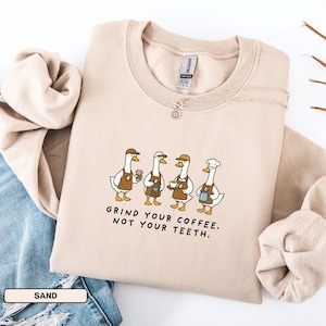 May include: A sand-coloured sweatshirt featuring a graphic of four geese dressed as baristas, each holding coffee-related items. The text "GRIND YOUR COFFEE. NOT YOUR TEETH." is printed below. A silver necklace is visible.