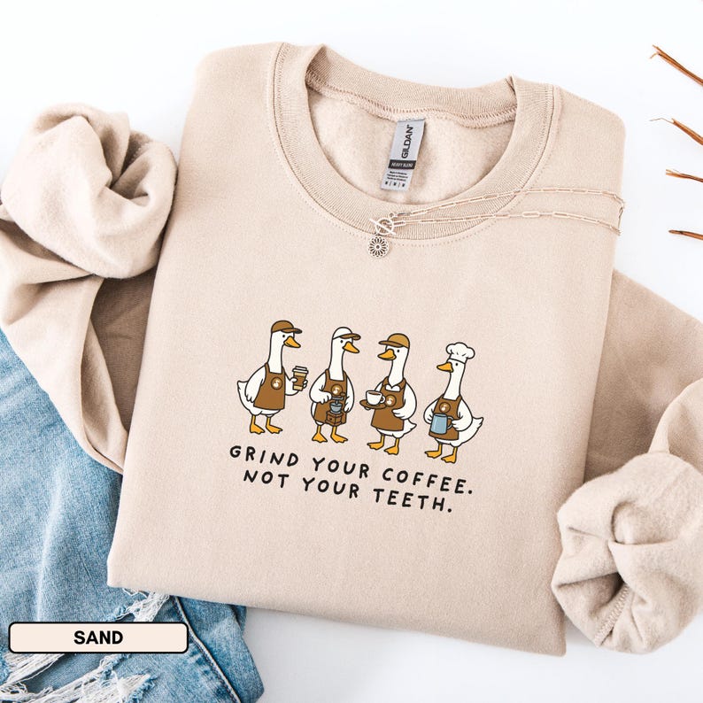 May include: A sand-coloured sweatshirt featuring a graphic of four geese dressed as baristas, each holding coffee-related items. The text "GRIND YOUR COFFEE. NOT YOUR TEETH." is printed below. A silver necklace is visible.
