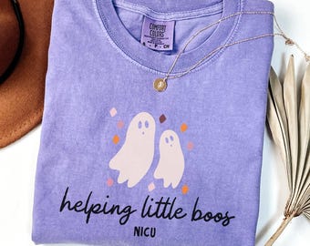以下が含まれることがあります： バイオレットカラーの半袖クルーネックTシャツ。グラフィックには、小さなオレンジとピンクのひし形に囲まれた2体の薄いピンクの幽霊が描かれています。幽霊の下には、黒い文字で「helping little boos」と「NICU」と記されています。