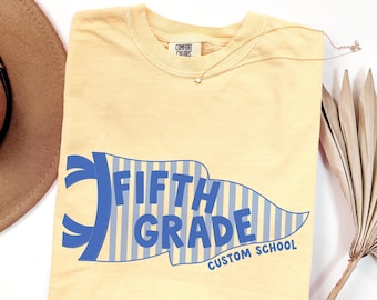 Peut inclure: Un t-shirt à manches courtes jaune beurre avec un motif de fanion rayé bleu et blanc portant l'inscription FIFTH GRADE et CUSTOM SCHOOL en lettres bleues. Le t-shirt présente une étiquette de marque Comfort Colors à l'encolure.