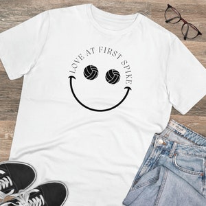 Puede incluir: Camiseta blanca con un diseño de cara sonriente negra. Los ojos de la cara sonriente son pelotas de voleibol. El texto "Love at First Spike" está escrito alrededor de la cara sonriente.