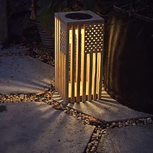 May include: A wooden outdoor lantern with a square top and a design resembling the American flag. The lantern has vertical wooden slats and a section with star-shaped cutouts. The interior glows with warm light, creating a patriotic ambiance.