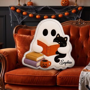 May include: A white ghost-shaped throw pillow featuring a black cat reading an orange book. The pillow includes a stack of two books in yellow and purple, a small orange jack-o'-lantern mug, and the name Sophia printed in black script lettering.