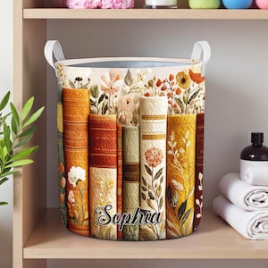 May include: A round laundry hamper with a floral and book design. The hamper features a cream-colored interior and white handles. The word "Sophia" is printed on the front in a script font. The design includes various colors such as orange, yellow, and green.
