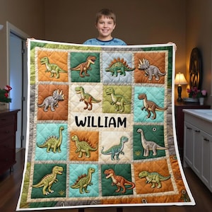Puede incluir: Una colcha colorida con ilustraciones de dinosaurios en un diseño de patchwork. La colcha tiene el nombre personalizado "WILLIAM" en el centro. Los cuadrados son en tonos de verde, naranja y azul, con un borde blanco.