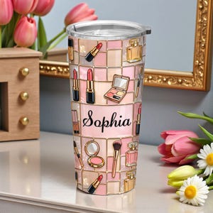 Op de afbeelding: Een gepersonaliseerde beker met een roze en gouden mozaïekpatroon, versierd met make-up illustraties. Het ontwerp bevat lippenstiften, parfumflesjes en make-up paletten. De naam "Sophia" staat in script op de voorkant.