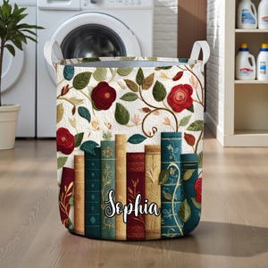 May include: A rectangular laundry hamper with white handles. The hamper features a floral design with red roses, green leaves, and a row of books. The name "Sophia" is written on the front in cursive.