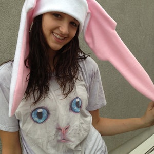 May include: A white fleece hat with long pink bunny ears and a red circle on the top. The hat is worn by a person with long brown hair and a white t-shirt with a cat graphic.