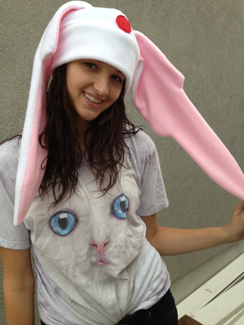 May include: A white fleece hat with long pink bunny ears and a red circle on the top. The hat is worn by a person with long brown hair and a white t-shirt with a cat graphic.