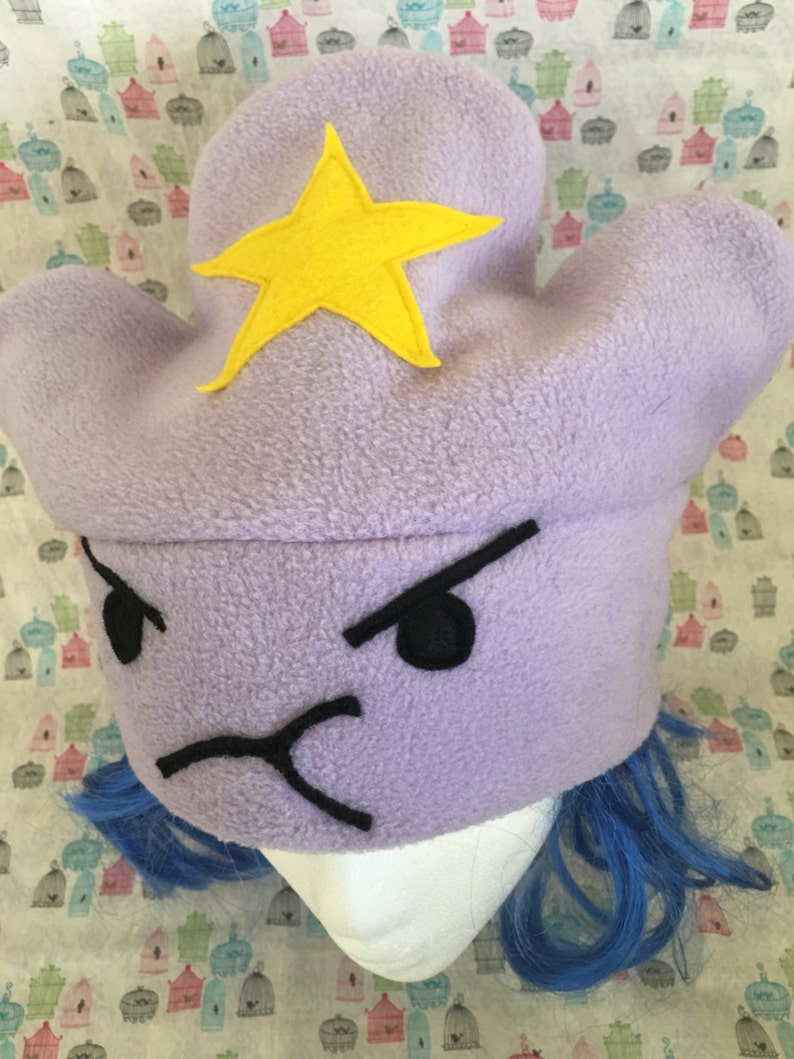 May include: Purple fleece Lumpy Space Princess hat with a yellow felt star and embroidered black facial features. The hat is shown on a mannequin head with bright blue wig.