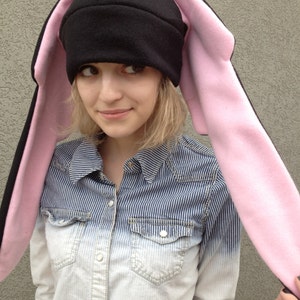 May include: A black fleece hat with long pink bunny ears and a blue felt accent on the top. The hat is worn by a person with a striped blue and white shirt.