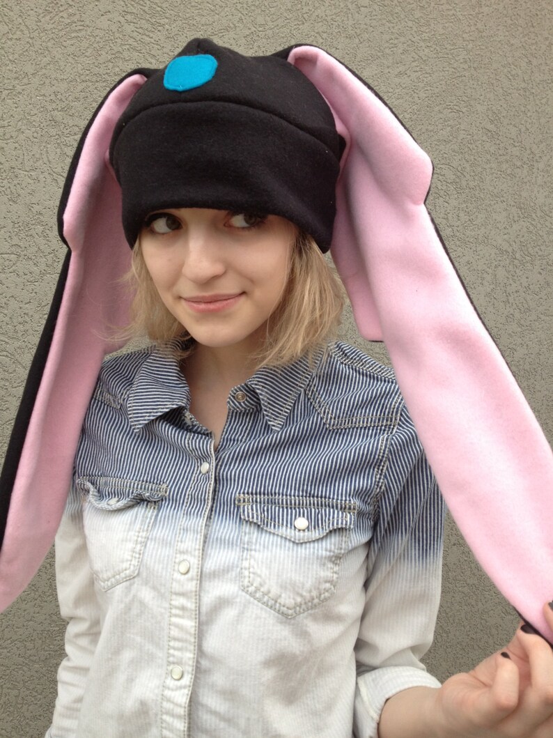 May include: A black fleece hat with long pink bunny ears and a blue felt accent on the top. The hat is worn by a person with a striped blue and white shirt.