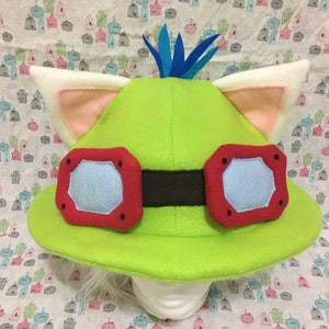 May include: Green and white plush hat with red goggles and blue feather detail. The hat has cat ears and a wide brim.