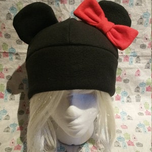 May include: Black fleece hat with large, rounded ears and a red bow on top. The hat is on a mannequin head with white hair.