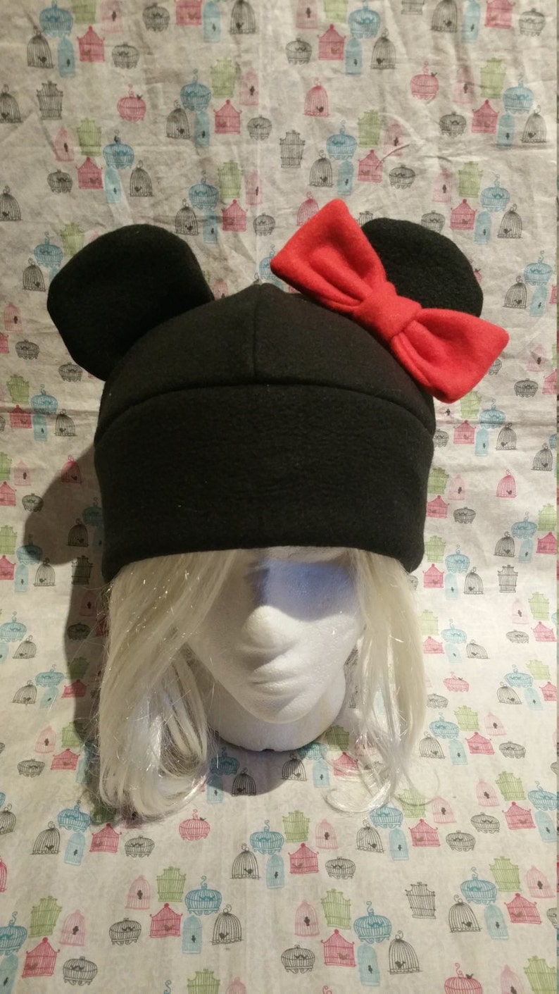 May include: Black fleece hat with large, rounded ears and a red bow on top. The hat is on a mannequin head with white hair.
