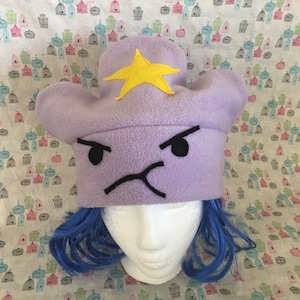 May include: Purple fleece Lumpy Space Princess hat with a yellow star. The hat features an embroidered frowning face with black details.  The hat is shown on a mannequin head with bright blue wig.