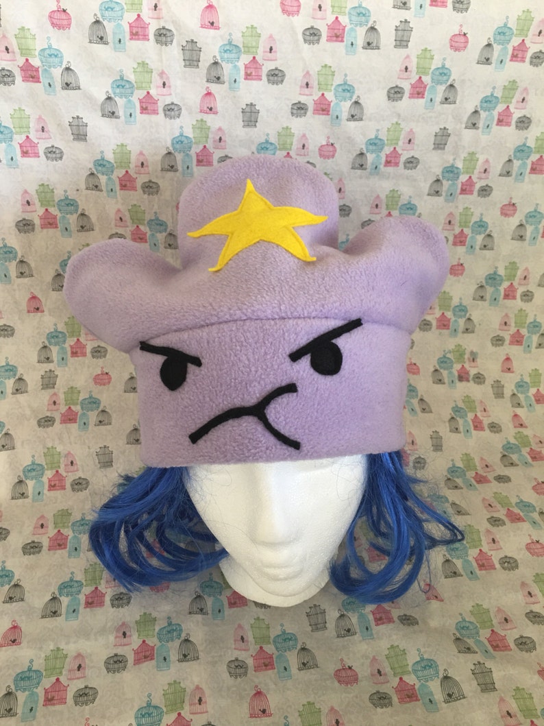 May include: Purple fleece Lumpy Space Princess hat with a yellow star. The hat features an embroidered frowning face with black details.  The hat is shown on a mannequin head with bright blue wig.