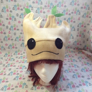 May include: A brown felt hat shaped like a tree with green felt leaves on top. The hat has a smiling face with black eyes and a brown nose.