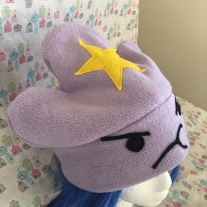 May include: Purple fleece Lumpy Space Princess hat with a yellow felt star and embroidered black facial features. The hat is shown on a mannequin head with blue wig.  The hat is a handmade, wearable costume accessory, perfect for fans of Adventure Time.