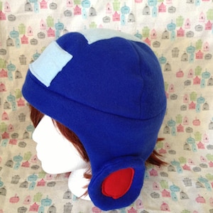 May include: A blue fleece hat with light blue and red accents. The hat features ear flaps and a light blue square patch on the top. The hat is designed to resemble a video game character.