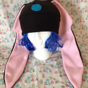 May include: Black fleece hat with long pink bunny ears. The hat has a blue circle on the top.