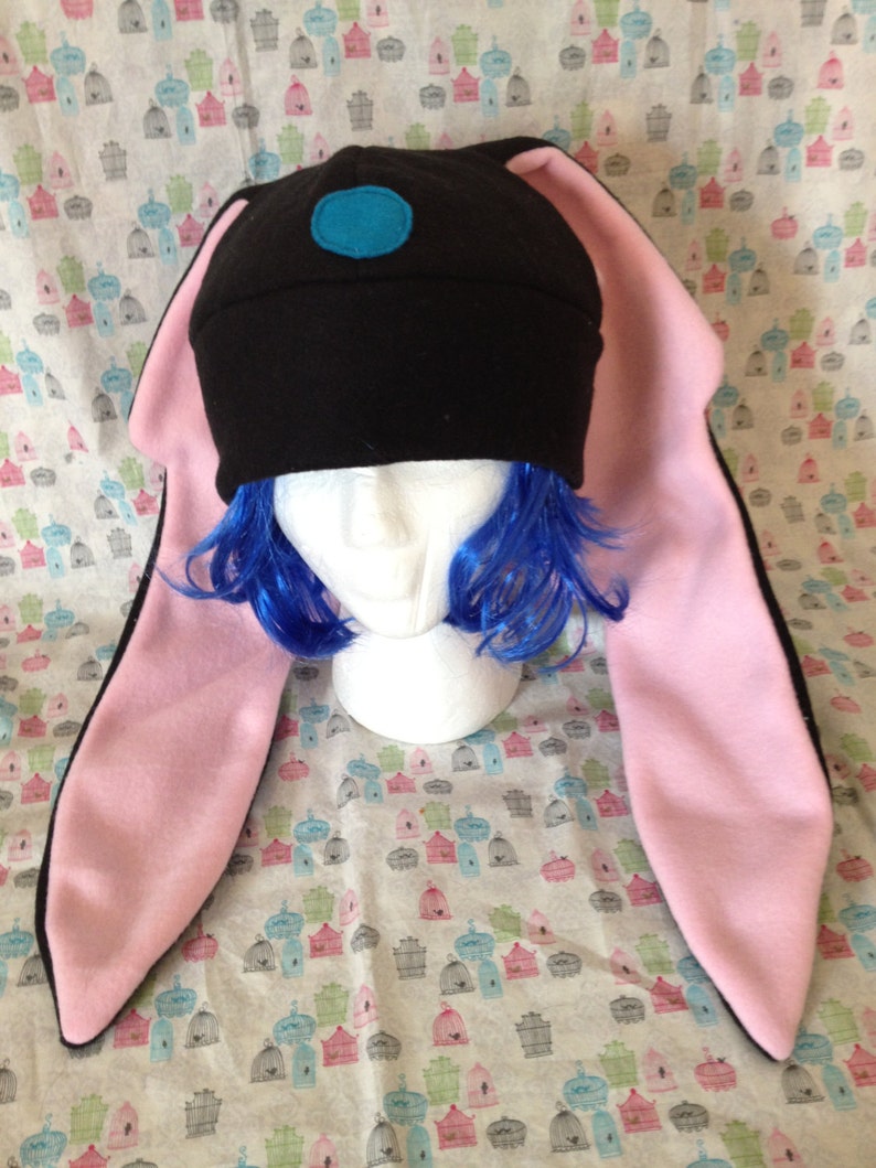 May include: Black fleece hat with long pink bunny ears. The hat has a blue circle on the top.