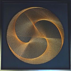 Op de afbeelding: Een ingelijst draadkunstwerk met een goud-oranje ontwerp tegen een donkerblauwe achtergrond. Het kunstwerk is samengesteld uit ingeweven draden, die een cirkelvormig, abstract patroon creëren. De lijst is van eenvoudig, zilverkleurig metaal.