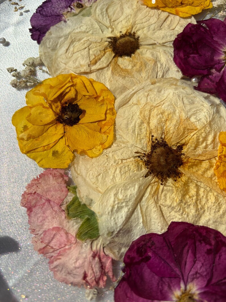 Puede incluir: Primer plano de flores secas en tonos amarillo, crema, rosa y morado. El arreglo presenta grandes flores texturizadas con centros oscuros y flores de acento m&aacute;s peque&ntilde;as. Los p&eacute;talos tienen una apariencia delicada y parecida al papel.