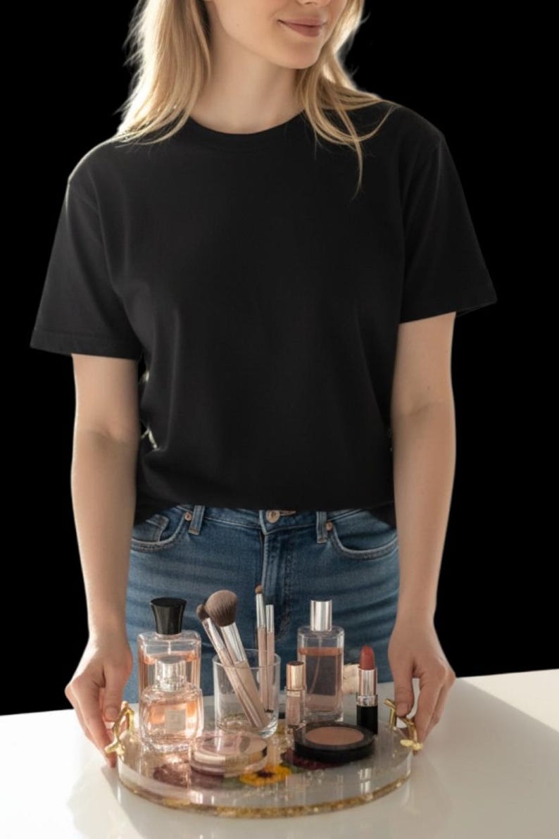 Puede incluir: Una camiseta negra de cuello redondo y vaqueros azules. Una bandeja redonda transparente con ribete dorado contiene frascos de perfume, brochas de maquillaje, l&aacute;piz labial y polveras. La bandeja est&aacute; decorada con flores.