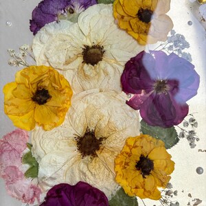 Puede incluir: Primer plano de un objeto redondo y transparente con flores secas. El arreglo incluye flores blancas, amarillas y moradas, con hojas verdes y peque&ntilde;as flores blancas. Copos de oro y peque&ntilde;as perlas est&aacute;n dispersos.