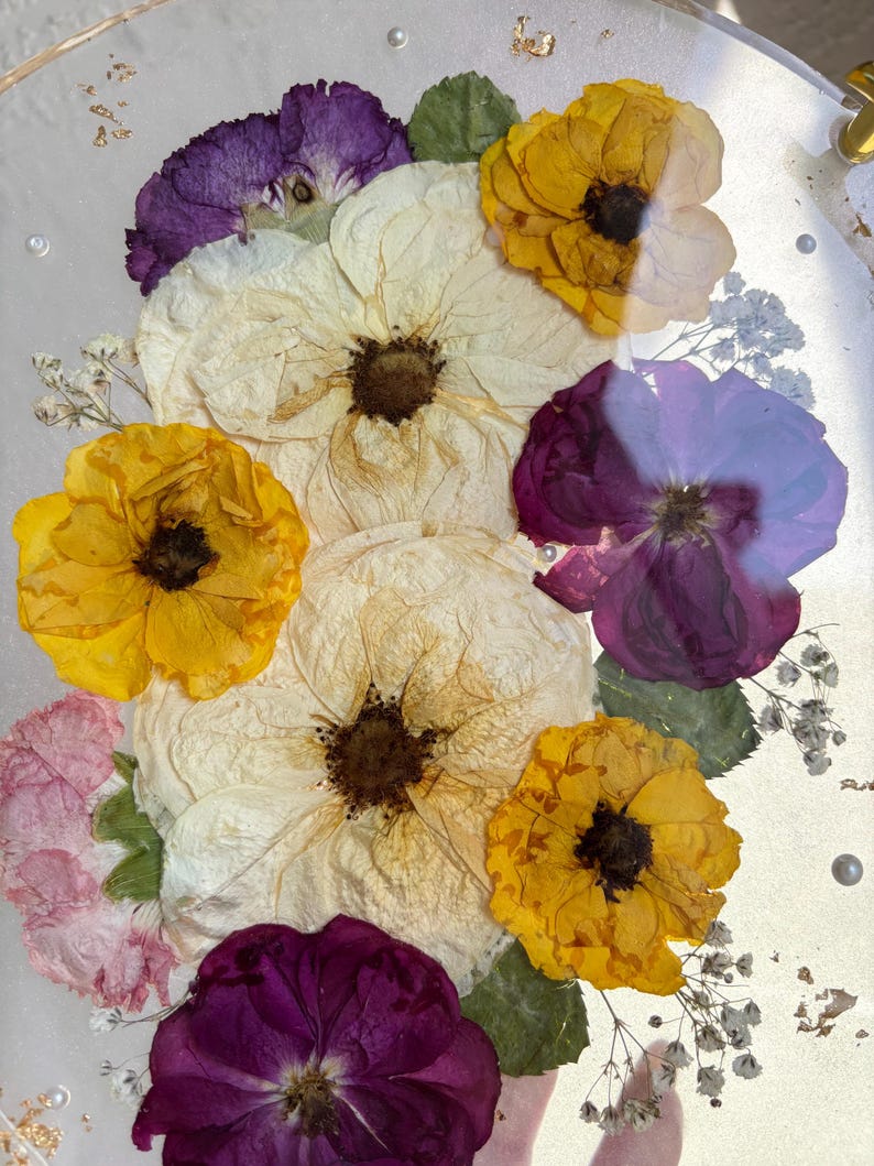 Puede incluir: Primer plano de un objeto redondo y transparente con flores secas. El arreglo incluye flores blancas, amarillas y moradas, con hojas verdes y peque&ntilde;as flores blancas. Copos de oro y peque&ntilde;as perlas est&aacute;n dispersos.
