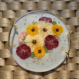 Puede incluir: Una bandeja decorativa redonda con una superficie azul claro, adornada con flores prensadas en tonos amarillos, crema y burdeos. La bandeja tiene asas doradas en forma de rama y est&aacute; adornada con peque&ntilde;as perlas y copos de oro.