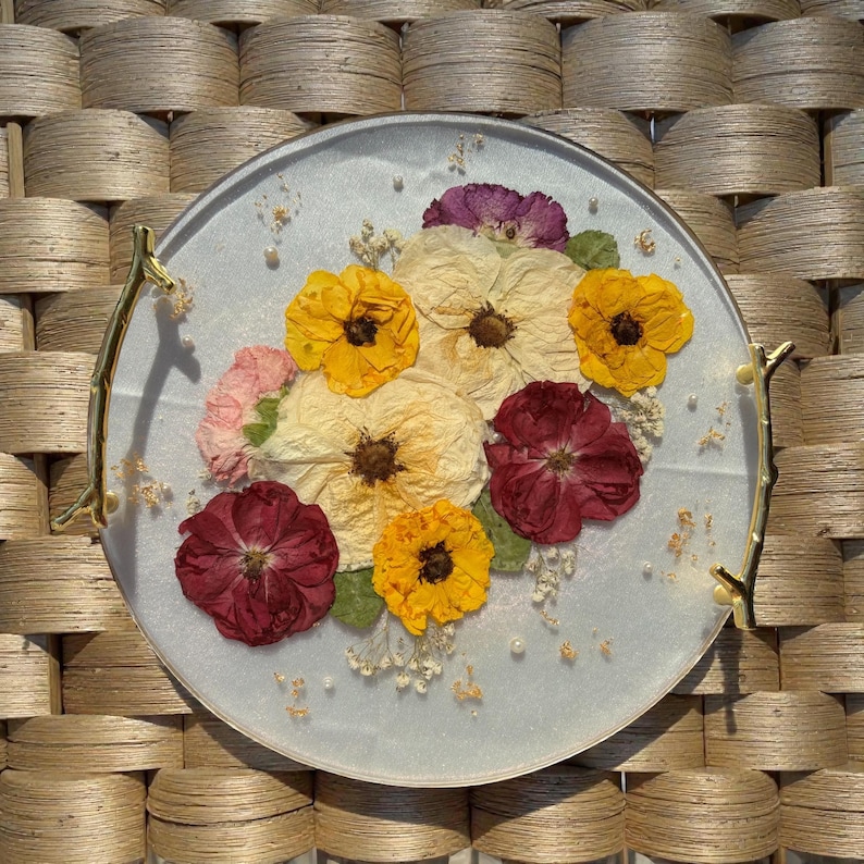 Puede incluir: Una bandeja decorativa redonda con una superficie azul claro, adornada con flores prensadas en tonos amarillos, crema y burdeos. La bandeja tiene asas doradas en forma de rama y est&aacute; adornada con peque&ntilde;as perlas y copos de oro.