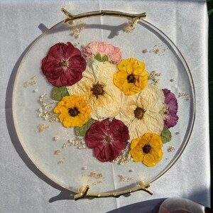 Puede incluir: Una bandeja redonda de resina transparente con flores secas en tonos rojos, amarillos, crema y morados. Las flores est&aacute;n dispuestas en un racimo y acentuadas con peque&ntilde;as perlas. La bandeja tiene asas doradas en forma de rama.