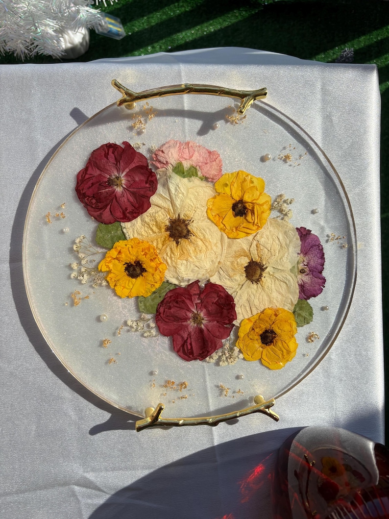 Puede incluir: Una bandeja redonda de resina transparente con flores secas en tonos rojos, amarillos, crema y morados. Las flores est&aacute;n dispuestas en un racimo y acentuadas con peque&ntilde;as perlas. La bandeja tiene asas doradas en forma de rama.