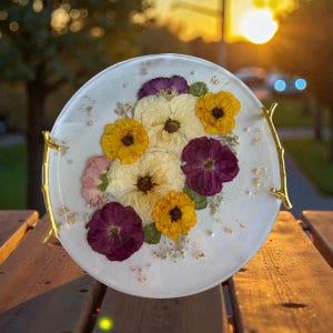 Puede incluir: Una bandeja redonda de resina blanca con flores secas incrustadas en tonos amarillos, morados y crema. La bandeja tiene asas doradas en forma de rama y est&aacute; decorada con copos de pan de oro y peque&ntilde;as perlas blancas.