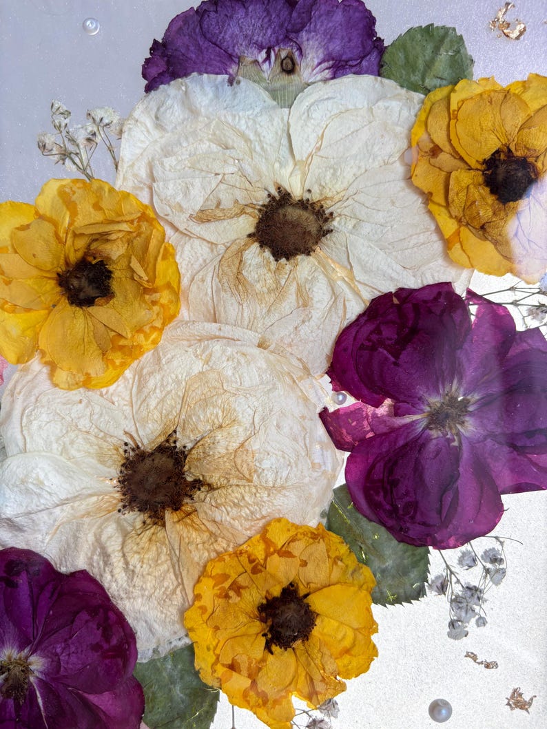 Puede incluir: Arreglo floral enmarcado con flores prensadas en tonos blancos, amarillos y morados. El arreglo incluye grandes flores abiertas y flores de acento m&aacute;s peque&ntilde;as, sobre un fondo claro con elementos decorativos.