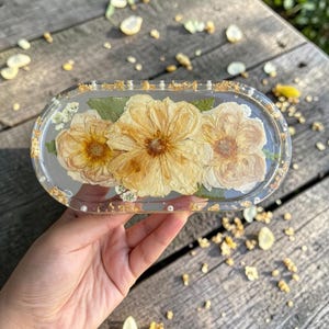 May include: An oval-shaped clear resin tray featuring three pressed yellow flowers and green leaves embedded within the material. The edges of the tray are decorated with small white beads and gold leaf flakes, creating a textured border.