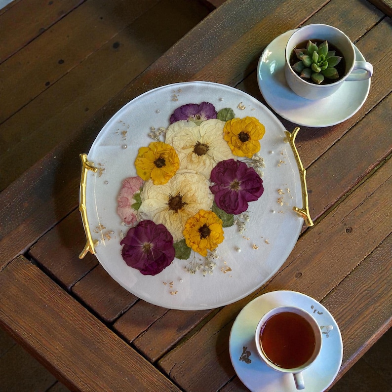 Puede incluir: Una bandeja redonda y blanca con asas doradas, adornada con flores secas amarillas, moradas y crema. La bandeja est&aacute; decorada con detalles de pan de oro y peque&ntilde;as perlas. Una taza de t&eacute; con t&eacute; y una taza con una suculenta est&aacute;n sobre una superficie de madera.