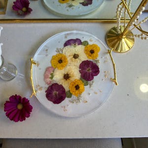 Puede incluir: Una bandeja redonda de resina transparente con flores secas en tonos amarillos, morados y crema. La bandeja tiene asas doradas y peque&ntilde;as l&aacute;minas doradas incrustadas. Una flor magenta est&aacute; cerca.