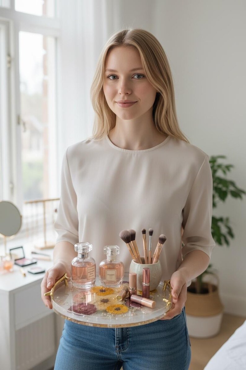 Puede incluir: Una mujer sostiene una bandeja decorativa con productos de belleza. La bandeja contiene dos frascos de perfume, brochas de maquillaje, barras de labios y flores secas. La bandeja tiene asas doradas y una superficie transparente. La mujer lleva un top color crema y vaqueros azules.