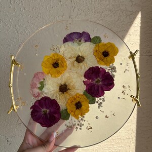 Puede incluir: Una bandeja redonda y transparente con asas doradas, incrustada con flores secas amarillas, moradas y blancas. La bandeja tiene un borde dorado y est&aacute; decorada con peque&ntilde;as escamas doradas y cuentas transparentes.