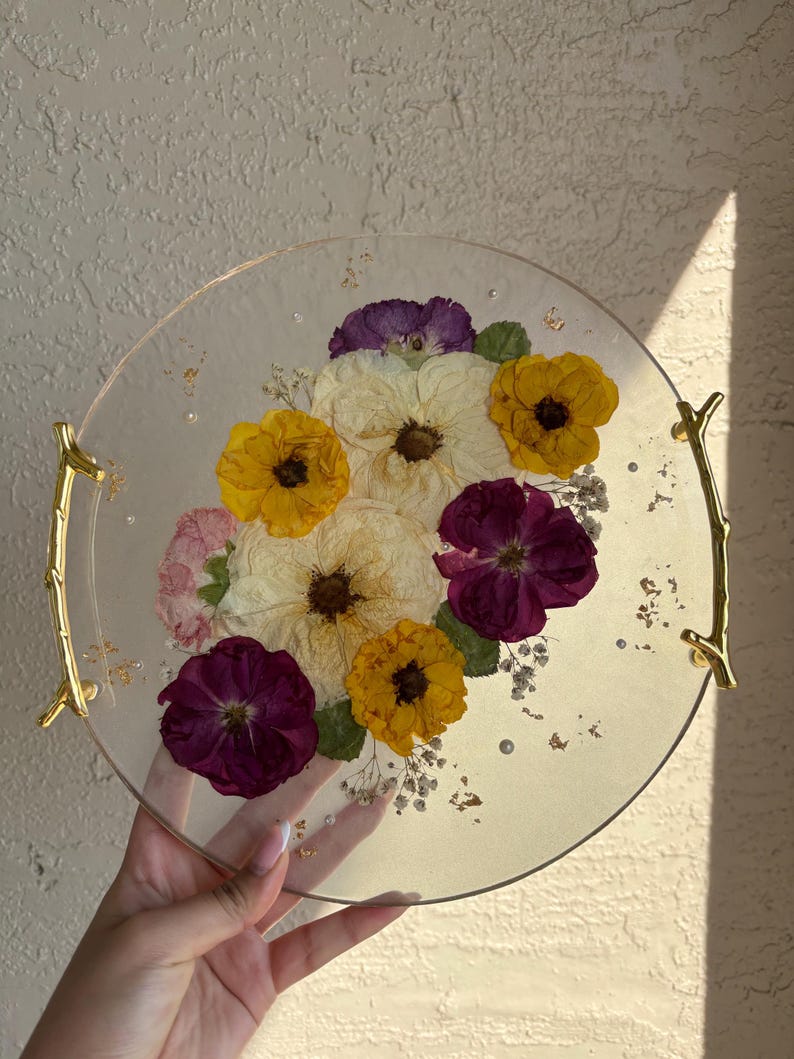Puede incluir: Una bandeja redonda y transparente con asas doradas, incrustada con flores secas amarillas, moradas y blancas. La bandeja tiene un borde dorado y est&aacute; decorada con peque&ntilde;as escamas doradas y cuentas transparentes.