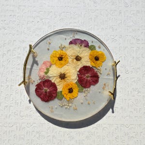 Puede incluir: Una bandeja redonda de resina transparente con flores secas en tonos amarillos, crema, rojos y morados. La bandeja tiene asas doradas y est&aacute; decorada con peque&ntilde;as escamas doradas y cuentas blancas. Las flores est&aacute;n dispuestas en un ramo.