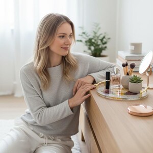 Puede incluir: Una c&oacute;moda de madera clara con una bandeja dorada que contiene frascos de perfume, brochas de maquillaje y una peque&ntilde;a suculenta. Un peque&ntilde;o espejo y un estuche compacto de oro rosa tambi&eacute;n est&aacute;n en la c&oacute;moda. Una mujer con un su&eacute;ter gris y pantalones blancos est&aacute; sentada cerca.