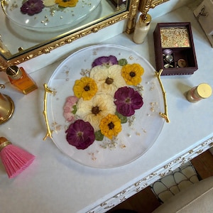 Puede incluir: Una bandeja redonda y transparente con asas doradas, adornada con flores secas amarillas, moradas y blancas. La bandeja est&aacute; decorada con detalles de pan de oro y peque&ntilde;as perlas, creando una pieza decorativa elegante.